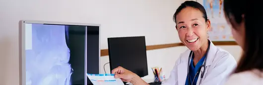 Foto de uma mulher olhando para o lado sorrindo enquanto segura uma folha de papel com informações escritas nessa folha. Ao lado dela há monitores com telas de raio-x.  A mulher é adulta, asiática, de cabelo escuro preso em rabo de cavalo, veste jaleco branco com uniforme hospitalar azul por debaixo. 