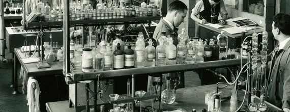 Fotografia antiga do departamento farmacêutico da Sandoz. Foto em preto e branco de uma sala estilo laboratório. Algumas pessoas vestida de jaleco e roupa social estão trabalhando com fracos de vidro dispostos numa mesa de dois níveis, no nível superior existem vários fracos de vidro e na mesa de baixo equipamentos e espaço livre. 