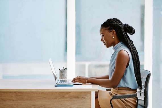 Foto de uma mulher sentada à mesa com as mãos no notebook, vista de lado. A mulher é negra, de cabelo longo com tranças, amarrado num coque. Ela veste regata de gola alta azul e saia bege. Ela está numa sala com iluminação clara. 
