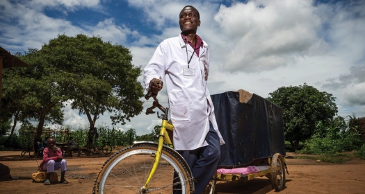 HCP wearing a medical coat pulling a cart bicycle in rural african village