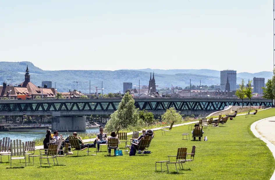 A panoramic view of Basel, from the Novartis Campus with people lounging in the foreground and the black forest in the background